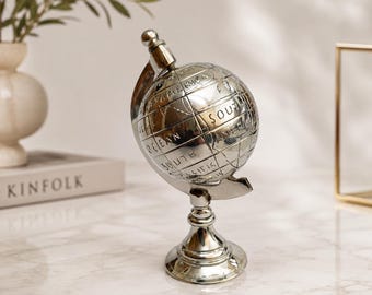 Może przedstawiać: Ozdobny globus z polerowanego metalu w kolorze srebrnym z wygrawerowanymi liniami geograficznymi i napisami, takimi jak South America i Pacific Ocean. Sferyczny globus jest osadzony na pasującej srebrnej podstawie z zakrzywionym ramieniem.