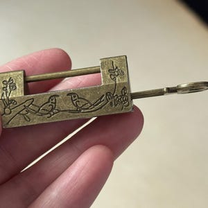 Puede incluir: Un candado de latón antiguo con una llave. El candado presenta un diseño grabado de dos pájaros posados en una rama con flores. La llave está insertada en la cerradura. El metal tiene un aspecto ligeramente envejecido.
