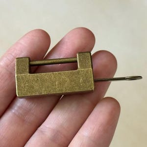 Puede incluir: Un pequeño candado rectangular de latón con una llave insertada. El candado tiene un diseño simple con una barra horizontal y una cerradura. La llave es delgada y tiene un extremo circular. El candado se sostiene en una mano.