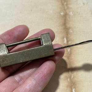 Puede incluir: Un candado rectangular de metal en tono latón con un arco horizontal y una llave metálica plana y delgada insertada en el lateral. El candado presenta un acabado desgastado de estilo antiguo con una textura metálica visible.