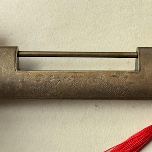 Puede incluir: Un candado rectangular de latón con un diseño grabado de dos peces koi flanqueando una flor de loto central. El metal tiene un acabado antiguo y desgastado. Una borla decorativa de color rojo brillante está unida al mecanismo del candado.