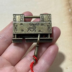 Puede incluir: Un candado de latón antiguo con intrincados diseños grabados. El candado presenta un cuerpo rectangular con patrones decorativos y una barra horizontal. Se inserta una llave con una cuerda roja. La estética general es antigua.