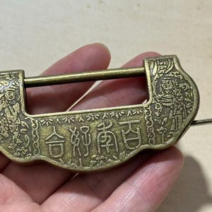 May include: Antique brass-colored lock with Chinese characters and detailed relief carvings. The lock features a rectangular body with a curved top and a sliding bar. Decorative figures are on the sides.