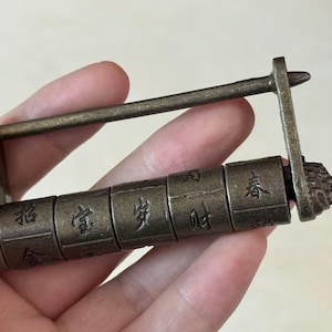 Puede incluir: Un candado de combinación de metal en tono bronce de forma rectangular con cinco cilindros giratorios. Cada cilindro está grabado con distintos caracteres chinos. El candado tiene un arco recto, largo y fino en la parte superior.