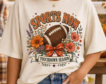 Puede incluir: Una camiseta de manga corta color hueso con un estampado gráfico que incluye el texto SPORTS MOM y TOUCHDOWN SEASON. El diseño presenta un balón de fútbol americano marrón, flores naranjas y rojas, hojas verdes y un lazo rojo central.