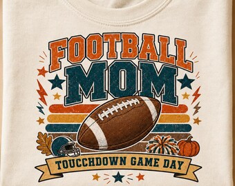 Puede incluir: Una camiseta de color crema con un gráfico que incluye el texto FOOTBALL MOM y TOUCHDOWN GAME DAY. El diseño presenta un balón de fútbol americano marrón, un casco azul, un pompón colorido, una calabaza naranja, estrellas y rayos.