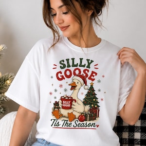 May include: A white short-sleeve t-shirt featuring a graphic of a goose wearing a Santa hat, holding a red mug labelled SILLY GOOSE, surrounded by a Christmas tree, presents, and snowflakes, with the text SILLY GOOSE Tis The Season printed above and below.