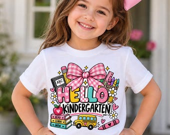 Puede incluir: Una camiseta blanca de manga corta con un estampado gráfico colorido que incluye un gran lazo rosa de cuadros, material escolar, un autobús escolar amarillo y el texto "HELLO KINDERGARTEN" en letras negritas y multicolores con iconos.