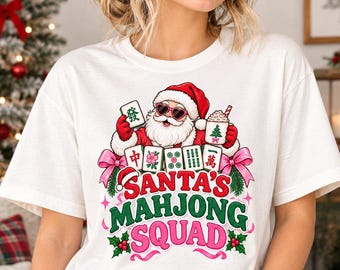Puede incluir: Una camiseta blanca con un gráfico de Santa Claus con gafas de sol en forma de corazón, sosteniendo una ficha de mahjong y una taza con un árbol de Navidad. El texto "SANTA'S MAHJONG SQUAD" aparece en rojo y verde, rodeado de fichas y lazos rosas.