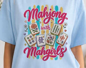 Puede incluir: Una camiseta azul claro con un estampado gráfico que incluye el texto "Mahjong with Mahgirls" en letra cursiva rosa. El diseño presenta cinco fichas de mahjong decoradas con motivos florales, de aves y caracteres, rodeadas de corazones, estrellas y flores.
