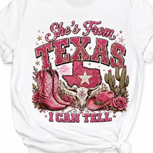 May include: A white t-shirt featuring a graphic print with the text "She's From TEXAS I CAN TELL". The design includes a pink leopard-print Texas state map, pink cowboy boots, a cow skull, a pink cowboy hat, cacti, and pink roses.