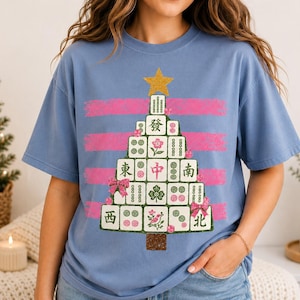 May include: A light blue t-shirt featuring a graphic of a Christmas tree constructed from mahjong tiles. The tree is topped with a gold star and decorated with pink bows and floral accents, set against horizontal pink brushstroke stripes.