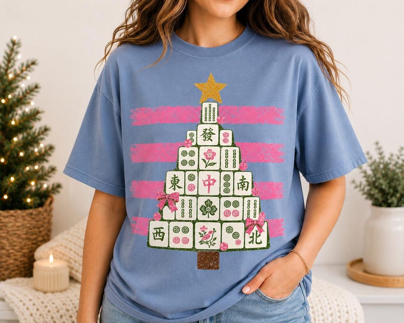 May include: A light blue t-shirt featuring a graphic of a Christmas tree constructed from mahjong tiles. The tree is topped with a gold star and decorated with pink bows and floral accents, set against horizontal pink brushstroke stripes.