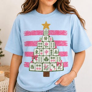 May include: A light blue t-shirt featuring a graphic of a Christmas tree constructed from Mahjong tiles. The tree is topped with a gold star and decorated with pink bows and ornaments, set against a background of horizontal pink brush strokes.