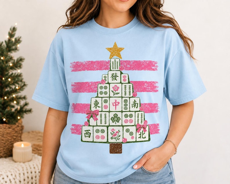 May include: A light blue t-shirt featuring a graphic of a Christmas tree constructed from Mahjong tiles. The tree is topped with a gold star and decorated with pink bows and ornaments, set against a background of horizontal pink brush strokes.
