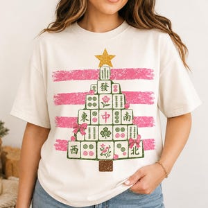 May include: A cream-coloured short-sleeve t-shirt featuring a graphic of a Christmas tree constructed from Mahjong tiles. The tree is topped with a gold star and decorated with pink bows, set against a background of horizontal pink brush strokes.
