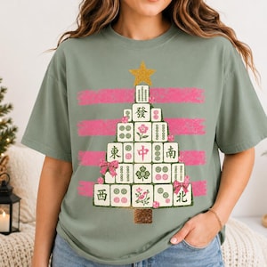 May include: A sage green short-sleeve t-shirt featuring a graphic of a Christmas tree constructed from Mahjong tiles. The tree is topped with a gold star and decorated with pink bows, set against horizontal pink brushstroke stripes.