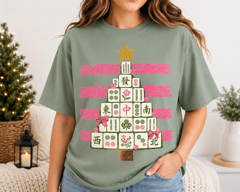 May include: A sage green short-sleeve t-shirt featuring a graphic of a Christmas tree constructed from Mahjong tiles. The tree is topped with a gold star and decorated with pink bows, set against horizontal pink brushstroke stripes.