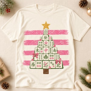 May include: A cream-coloured t-shirt featuring a graphic of a Christmas tree constructed from mahjong tiles. The tree is topped with a gold star and decorated with pink bows and small pink circles, set against horizontal pink brushstroke stripes.