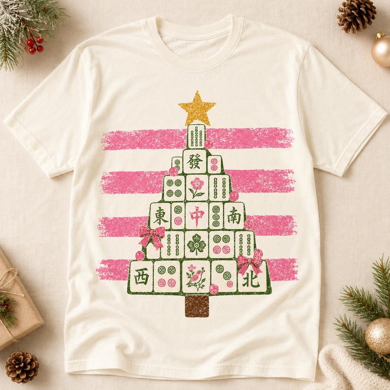 May include: A cream-coloured t-shirt featuring a graphic of a Christmas tree constructed from mahjong tiles. The tree is topped with a gold star and decorated with pink bows and small pink circles, set against horizontal pink brushstroke stripes.