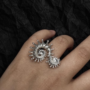 Könnte beinhalten: Ein silberfarbener Ring mit einem Spiraldesign, das eine Reihe spitzer Elemente aufweist. Der Ring wird an einem Finger getragen und zeigt seine komplizierten Details und seine einzigartige Form. Das Design des Rings erinnert an ein Nautilus-Gehäuse.