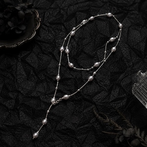 May include: A layered silver necklace featuring grey oval pearls. The necklace has a delicate chain and a Y-shaped design, with a single pearl pendant. The jewellery is displayed on a black textured surface.