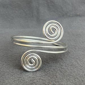 Pode incluir: Uma pulseira de braço prateada com dois desenhos em espiral. A pulseira tem um design de banda dupla que envolve o braço. As espirais estão localizadas na parte superior e inferior da pulseira.