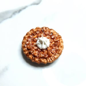May include: A miniature pie with a golden-brown crust and a filling of nuts and caramel. A dollop of white cream sits in the center. The pie is round and has a textured crust. The dessert is on a white surface.