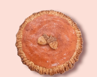 May include: A homemade pumpkin pie with a golden brown crust and a smooth, orange filling. Two decorative acorn shapes are placed on top of the pie.