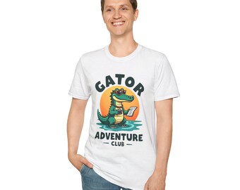 Puede incluir: Una camiseta blanca de manga corta con un gráfico de un caimán verde con gafas de sol y una cámara. El texto GATOR ADVENTURE CLUB está impreso en letras azul oscuro encima y debajo del caimán, que se encuentra frente a un sol naranja.