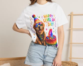 Puede incluir: Una camiseta blanca con el gráfico de un golden retriever que lleva un gorro de fiesta arcoíris y gafas de sol coloridas. El perro lleva un pañuelo que dice Level 4 Unlocked. Junto al perro hay un pastel de cumpleaños arcoíris con un 4.