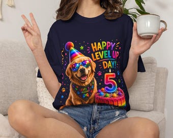 Puede incluir: Una camiseta azul marino oscuro con el gráfico de un golden retriever con un gorro de fiesta arcoíris y gafas de sol coloridas. El perro lleva un pañuelo que dice "LEVEL 5 UNLOCKED" junto a un pastel de cumpleaños con una vela del número 5 y el texto "HAPPY LEVEL UP DAY!"
