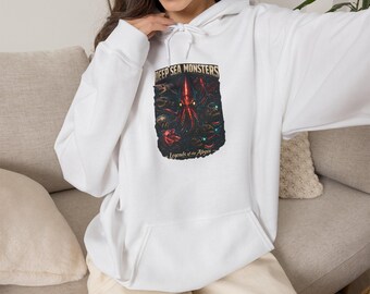 Puede incluir: Una sudadera con capucha blanca que presenta un estampado gráfico en el pecho. El diseño muestra una criatura roja similar a un calamar rodeada de vida marina oscura con el texto DEEP SEA MONSTERS y Legends of the Abyss en dorado.