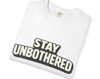 Puede incluir: Una camiseta blanca de manga corta con el texto STAY UNBOTHERED impreso en una fuente negra, audaz y desgastada, con un contorno de color crema. La prenda tiene un cuello redondo y una etiqueta interior visible que indica la talla L.