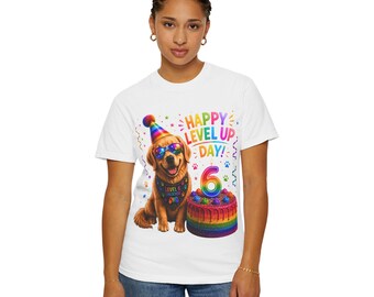 Puede incluir: Una camiseta blanca con el gráfico de un golden retriever con un gorro de fiesta y gafas de sol arcoíris, y un pañuelo que dice LEVEL 6 UNLOCKED. Junto al perro hay un pastel de cumpleaños de capas arcoíris con una vela del número 6 y el texto HAPPY LEVEL UP DAY!