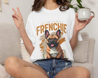 Puede incluir: Una camiseta blanca de manga corta con un gráfico de un bulldog francés marrón. El texto sobre el perro dice "LIFE IS BETTER WITH A FRENCHIE" en letras mayúsculas marrones. El diseño incluye un fondo de pincelada marrón y hierba verde.