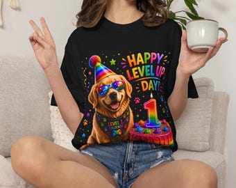Puede incluir: Una camiseta negra con un golden retriever que lleva un gorro de fiesta arcoíris y gafas de sol coloridas, un pañuelo con el texto 'LEVEL 1 UNLOCKED', un pastel de cumpleaños con una vela arcoíris y el texto 'HAPPY LEVEL UP DAY'.