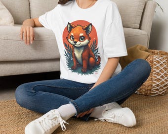 Puede incluir: Una camiseta blanca de manga corta con un estampado gráfico de un zorro de dibujos animados con ojos grandes, sentado entre follaje verde estilizado sobre un fondo circular naranja. El zorro tiene pelaje naranja y blanco y una cola tupida.