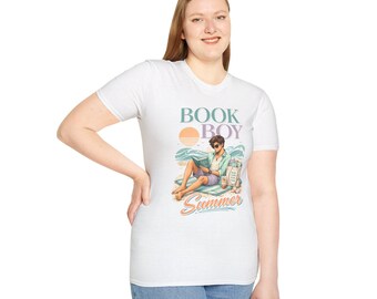 Puede incluir: Una camiseta blanca de manga corta con un estampado gráfico de una persona leyendo un libro en la playa. El texto dice BOOK BOY Summer en una fuente retro. La ilustración incluye un sol, olas del mar y una bolsa de tela con libros en la arena.