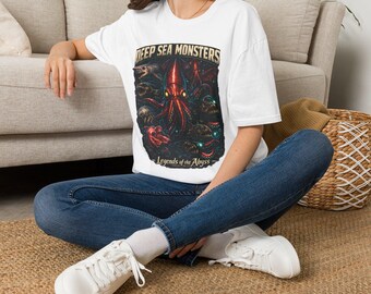 Puede incluir: Una camiseta blanca de manga corta con un estampado gráfico de varias criaturas marinas, incluido un gran calamar central con ojos brillantes. El texto DEEP SEA MONSTERS y Legends of the Abyss está impreso en una fuente de estilo vintage.