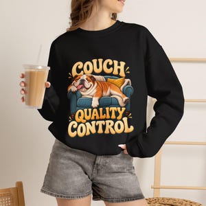 Könnte beinhalten: Ein schwarzes Langarm-Sweatshirt mit einer Grafik eines braun-weißen Bulldogs, der auf einem blauen Sofa schläft. Der Text "COUCH QUALITY CONTROL" ist in großen, gelben, abgerundeten Buchstaben über und unter der Hundeabbildung gedruckt.