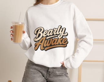 Puede incluir: Una sudadera blanca de manga larga con cuello redondo que presenta el texto Bearly Awake en una fuente retro de color marrón y negro. La sudadera se combina con pantalones cortos de mezclilla gris y se sostiene un vaso de plástico transparente.