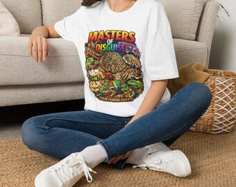 Puede incluir: Una camiseta blanca de manga corta con un estampado gráfico colorido de varios animales, incluyendo un camaleón, un pulpo y una rana. El texto MASTERS OF DISGUISE y COLOR CHANGING EXPERTS aparece en letras multicolores en el frente.
