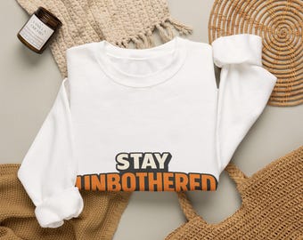 Puede incluir: Una sudadera blanca de manga larga con cuello redondo que presenta el texto STAY UNBOTHERED impreso en letras negritas y cuadradas. La palabra STAY es de color crema con un borde oscuro, mientras que la palabra UNBOTHERED es naranja vibrante.