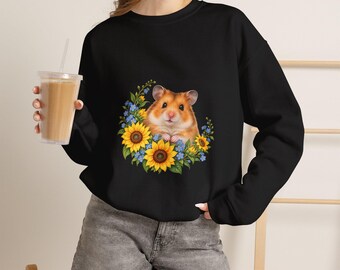 Puede incluir: Una sudadera negra de manga larga con un estampado gráfico de un hámster marrón rodeado de girasoles amarillos y pequeñas flores azules. El hámster está situado en el centro del arreglo floral con las patas recogidas.