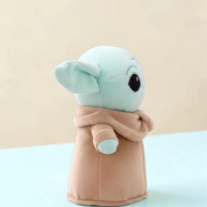 Puede incluir: Un peluche de un personaje con orejas grandes, cara verde claro y ojos negros grandes. El personaje lleva una t&uacute;nica beige. El juguete es un personaje popular de una franquicia de ciencia ficci&oacute;n.
