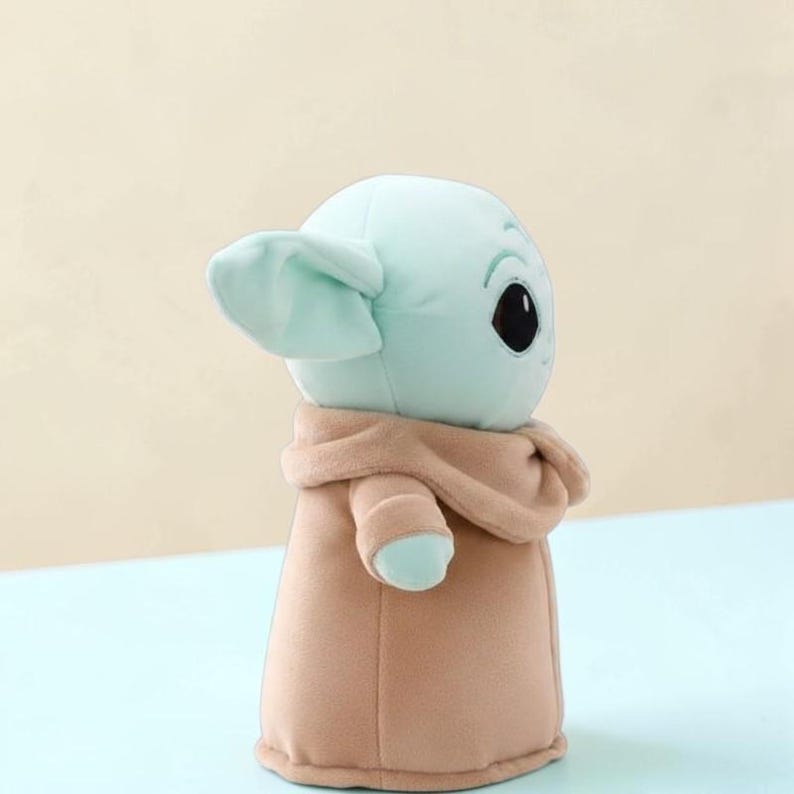 Puede incluir: Un peluche de un personaje con orejas grandes, cara verde claro y ojos negros grandes. El personaje lleva una t&uacute;nica beige. El juguete es un personaje popular de una franquicia de ciencia ficci&oacute;n.