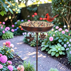 May include: A bronze bird feeder filled with birdseed, with a decorative hummingbird and flower design. A bright red cardinal perches on the edge. The feeder is set in a garden with a stone pathway and colourful flowers.