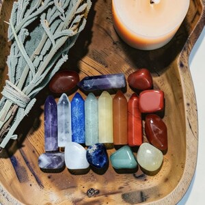 Könnte beinhalten: Eine Holzschale enthält eine Sammlung farbiger Kristalle, ein Salbeibündel und eine brennende Kerze. Die Kristalle sind in einem Regenbogenspektrum angeordnet, mit verschiedenen Formen und Größen. Der Salbei ist gebündelt.
