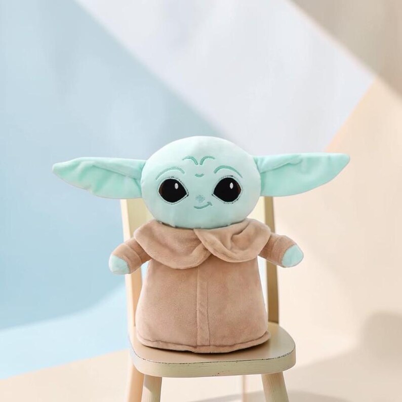 Puede incluir: Un mu&ntilde;eco de peluche de un personaje con orejas grandes, ojos negros grandes y tez verde claro. El mu&ntilde;eco lleva un abrigo color canela y est&aacute; sentado en una peque&ntilde;a silla de madera. El fondo es una suave mezcla de azul y beige.